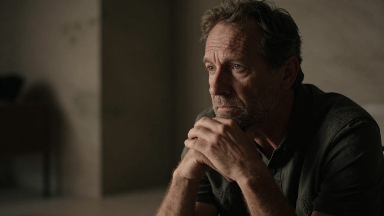 Hombre maduro con expresión reflexiva sentado en una habitación oscura, contemplando con serenidad en medio de la soledad.